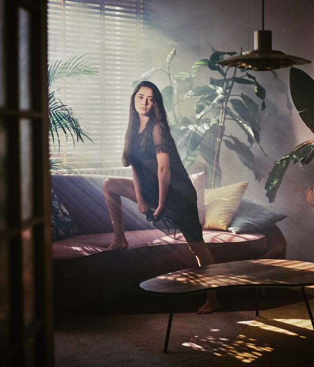 A woman in a sheer dress poses on a sofa in a room filled with plants and dramatic light rays.
