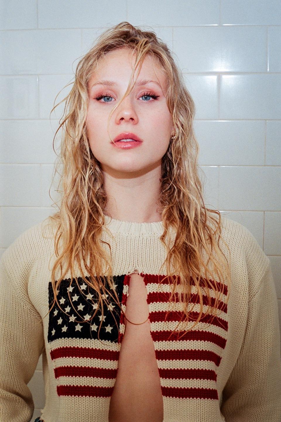 A woman with wet, wavy hair and pink eye makeup wears a sweater with an American flag design, revealing her chest.