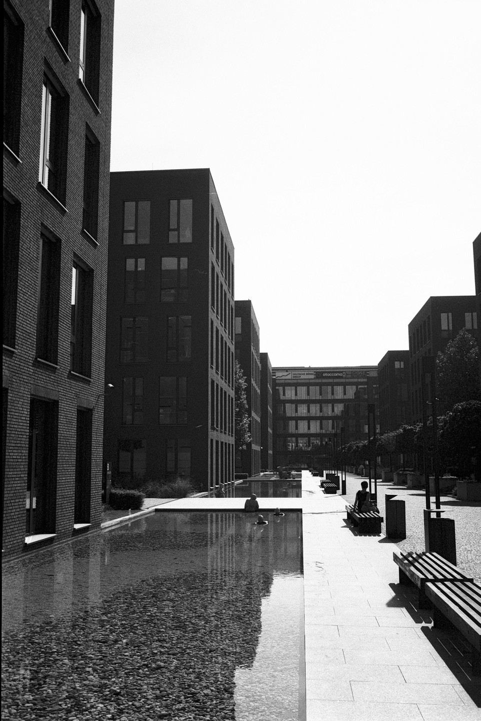 A person sits on a bench next to a long, narrow pool of water reflecting modern buildings under a bright sky.