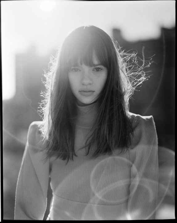 A woman with long hair and bangs looks directly at the viewer, backlit by the sun.