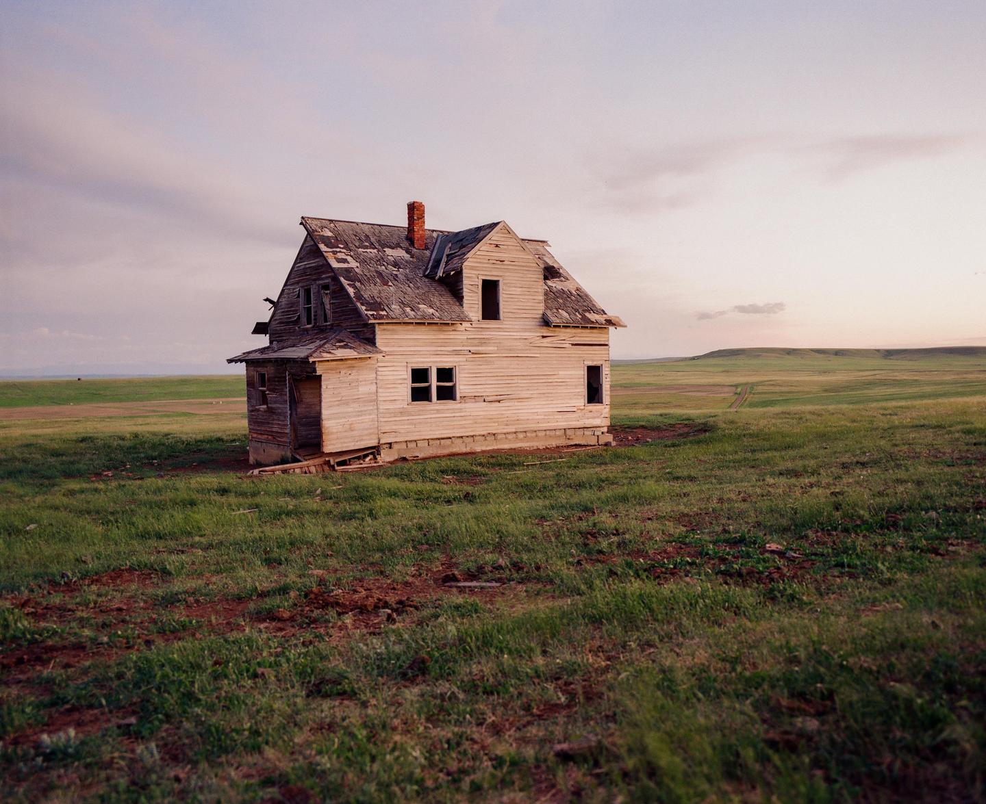 An abandoned house stands in a vast grassy field under a pale sky.