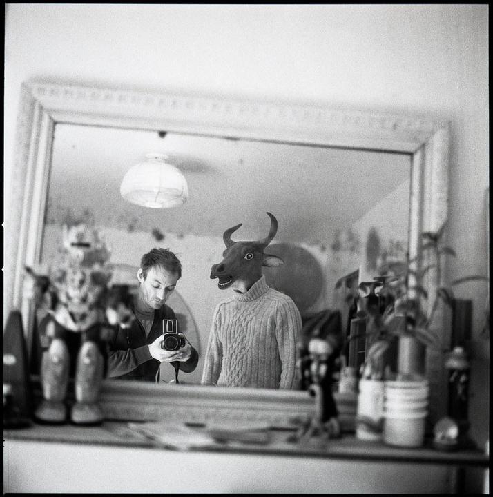 A man takes a mirror self-portrait next to a person wearing a cow mask and a sweater.