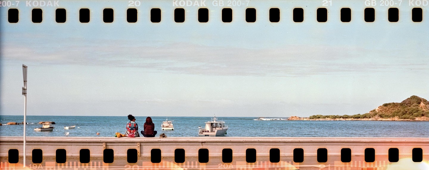 Two women sit on a concrete wall overlooking a body of water with boats and a distant island.