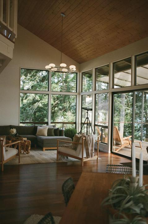 A modern living room with large windows overlooks a forest and a body of water.