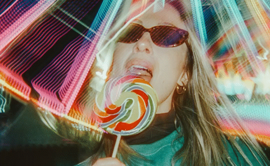 A woman with sunglasses licks a colorful lollipop against a blurred background of neon lights.
