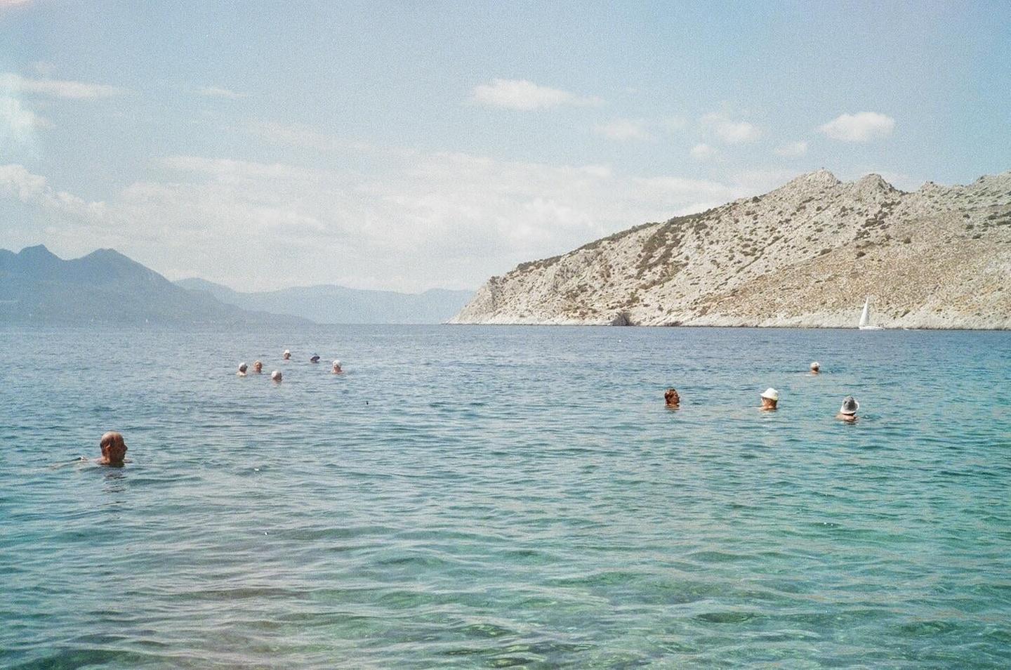 People swim in clear blue water with mountains and a sailboat in the background.