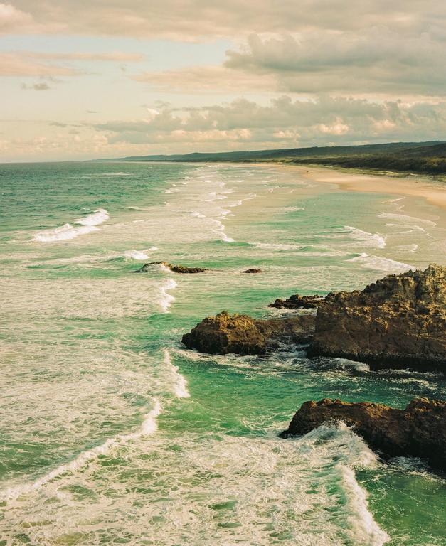 Waves crash against a rocky shore and a sandy beach stretches into the distance under a cloudy sky.
