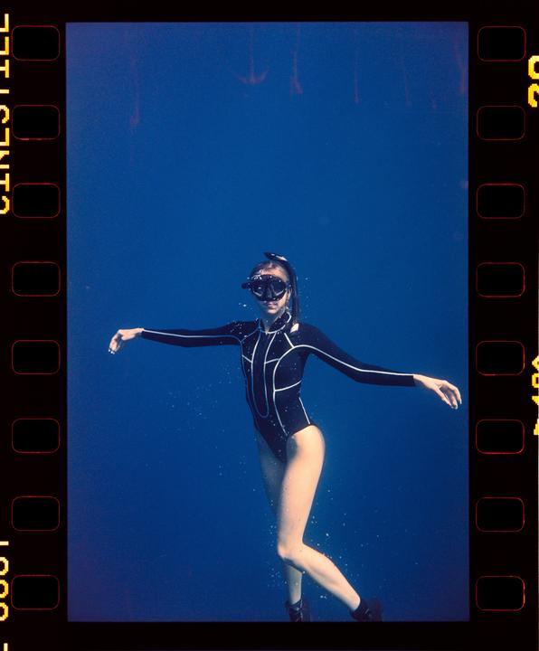 A woman in a black wetsuit and snorkel mask floats gracefully underwater in deep blue water.