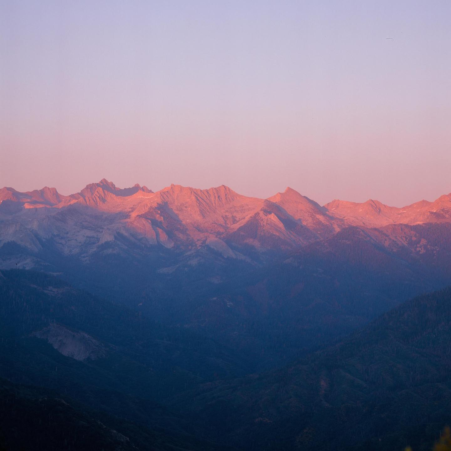 The sun illuminates the peaks of mountains above a dark valley.