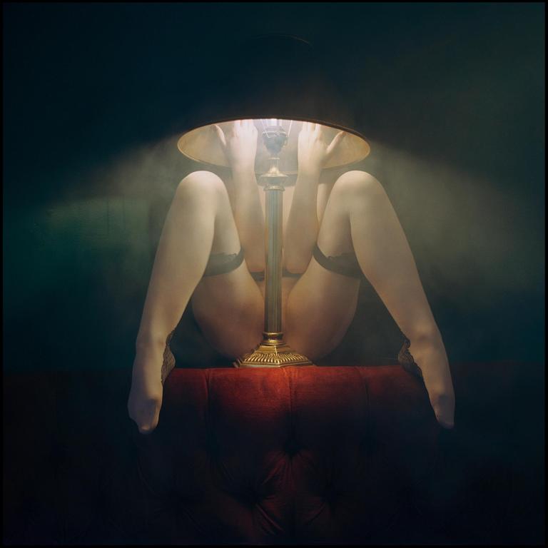 A woman's legs and hands are visible around a lit table lamp on a red tufted surface.