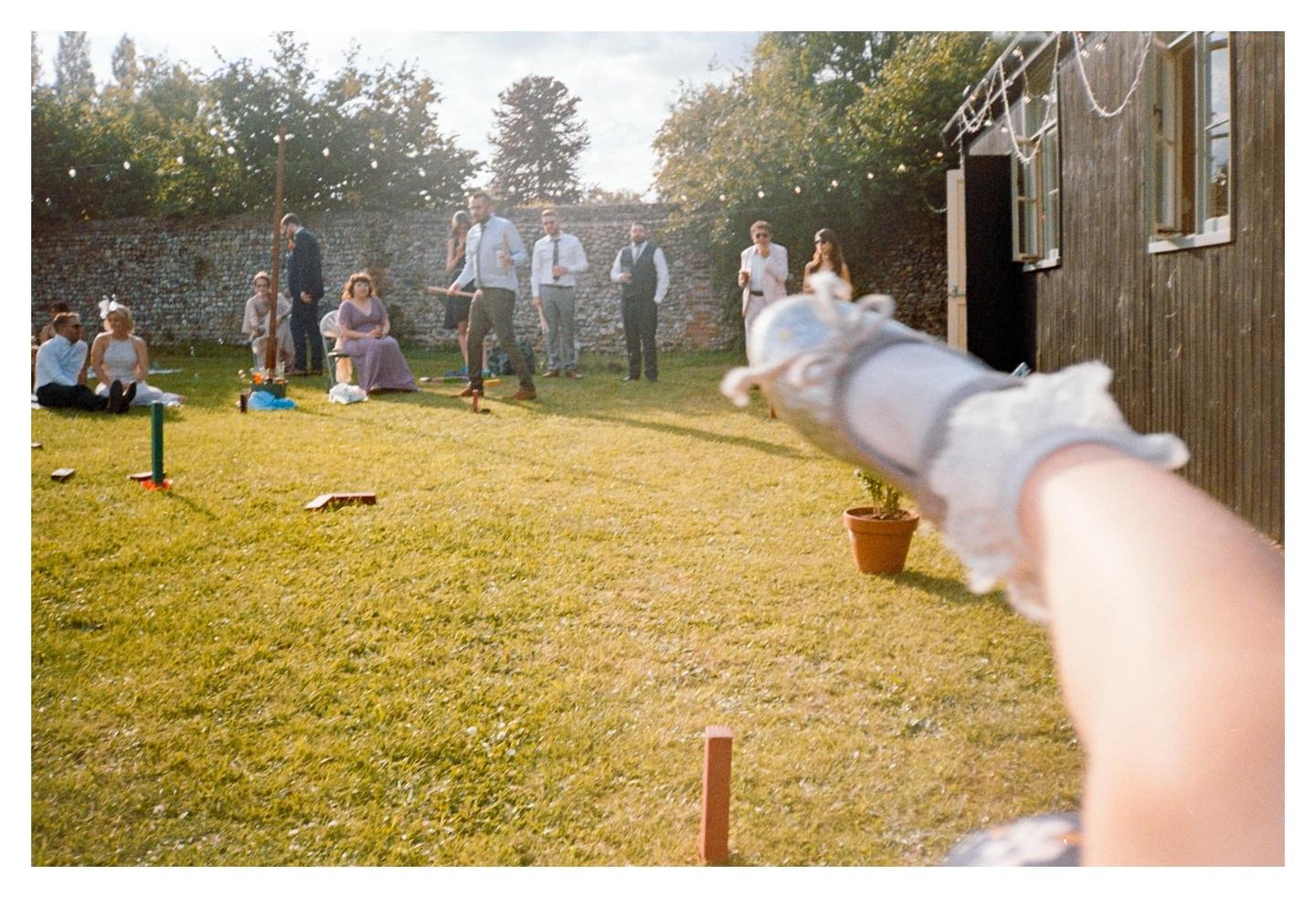 A child's arm holding a camera is visible in the foreground, pointed at a group of adults playing croquet on a sunny lawn.