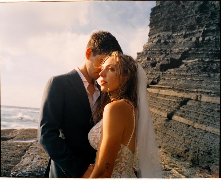 A couple stands on a rocky beach with a large rock formation behind them.