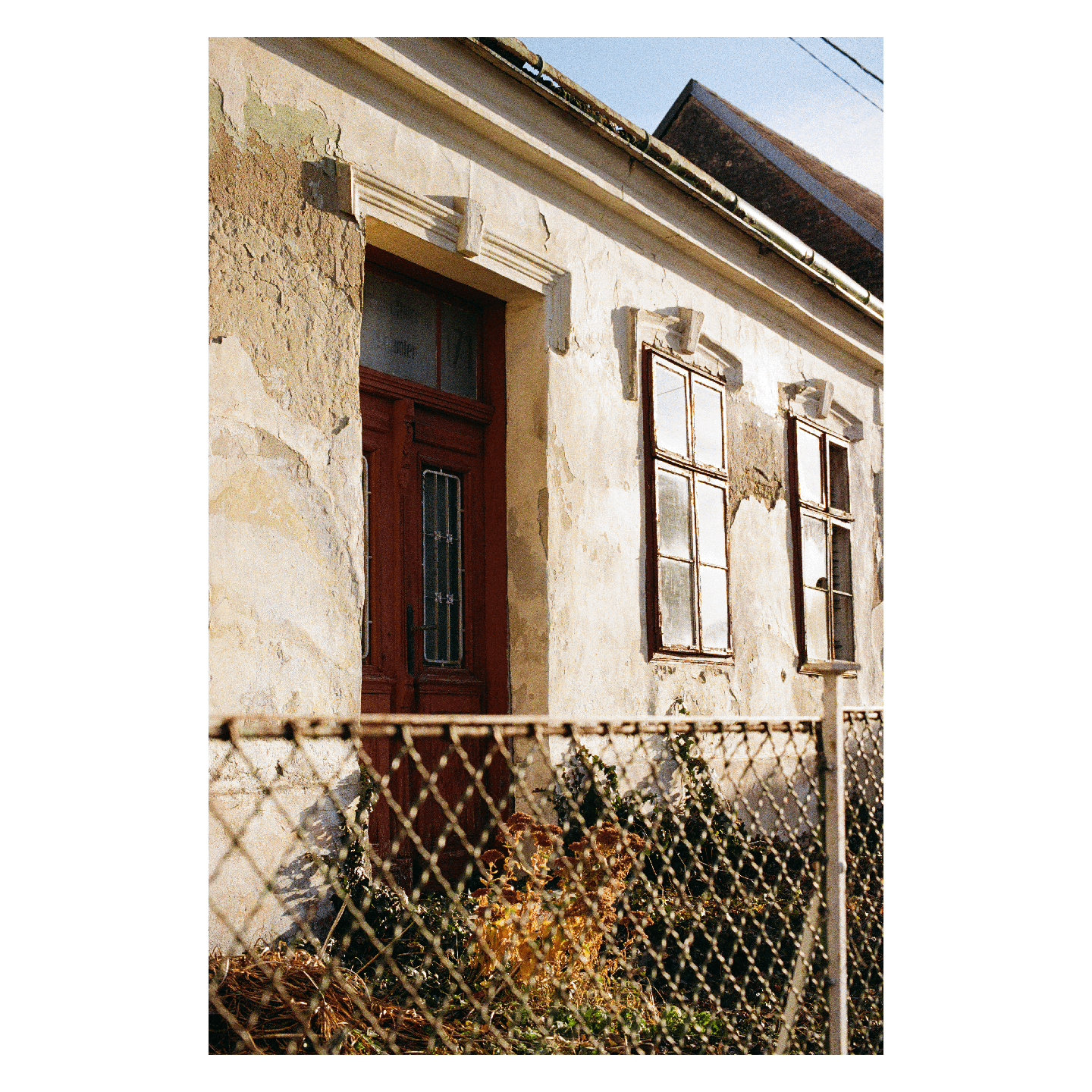 A weathered building with a red door and two windows is partially obscured by a chain link fence.