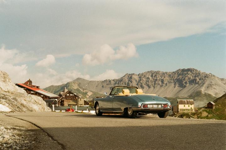 A light blue convertible car is parked on a winding road with mountains and buildings in the background.