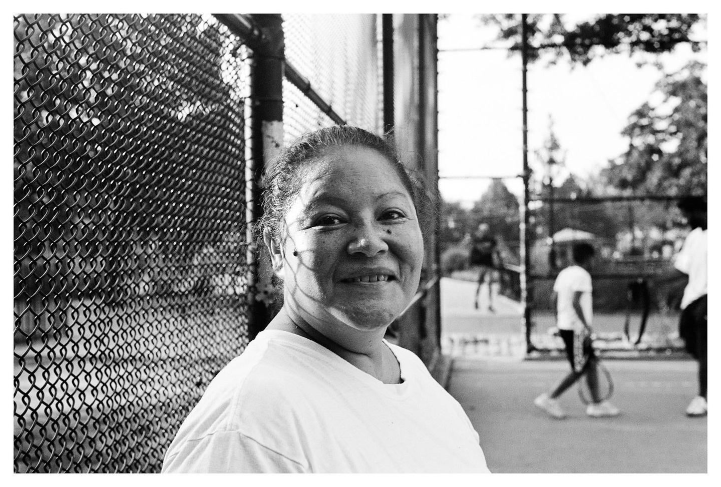 A woman smiles at the camera while standing next to a chain-link fence with people in the background.