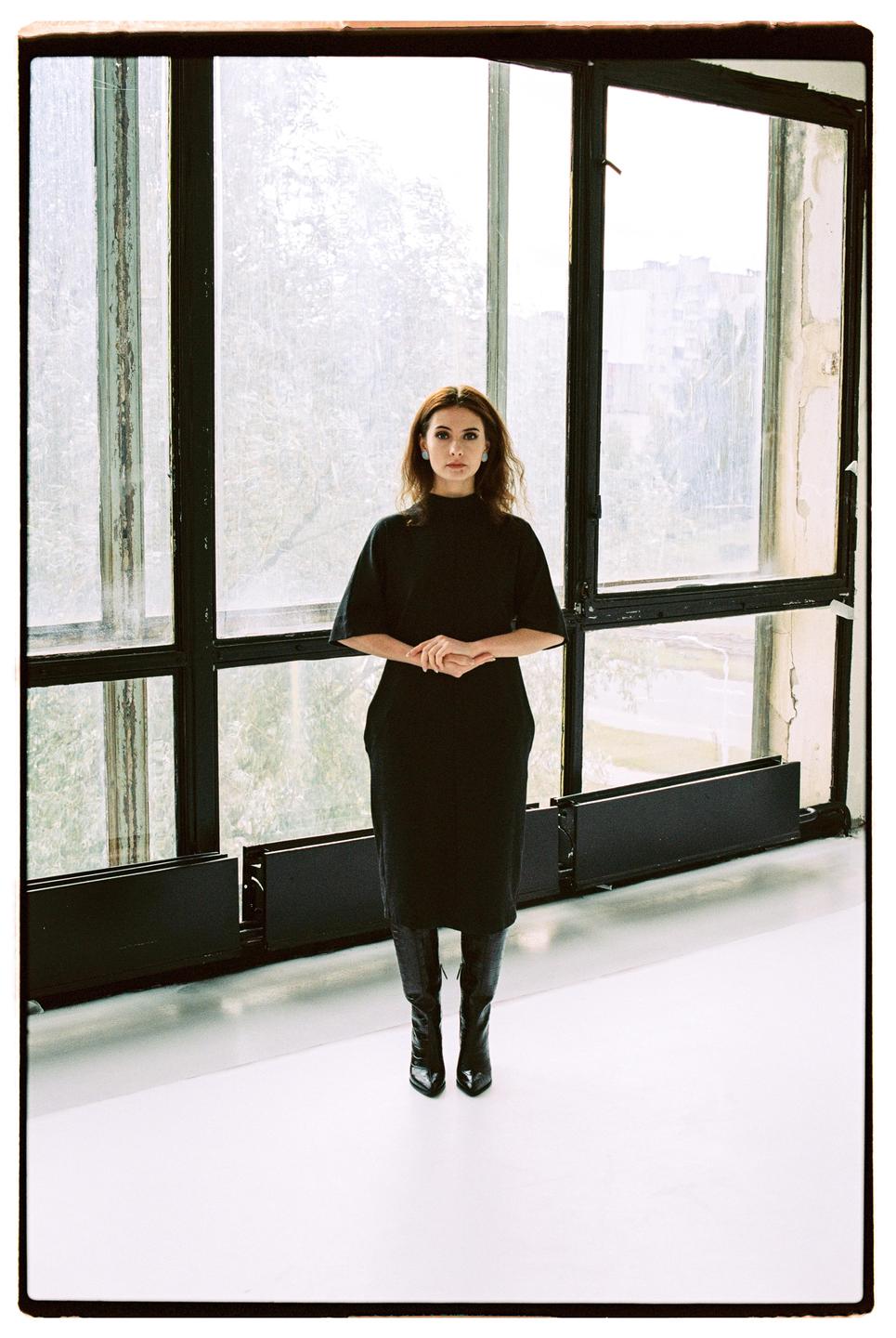 A woman in a black dress and boots stands in front of a large window.