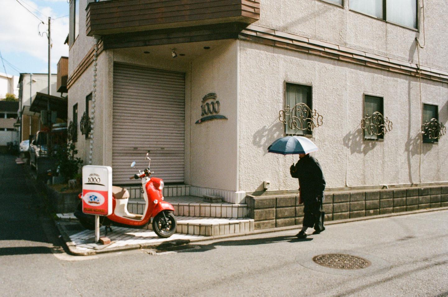 A person holding an umbrella walks past a red scooter parked on a street next to a building.