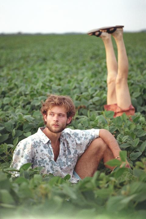 A man sits in a field of green plants while a pair of legs with sandals points upwards in the background.