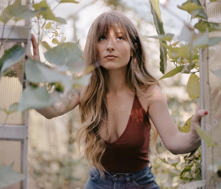 A woman with long hair and bangs looks up while standing in a greenhouse surrounded by green foliage.