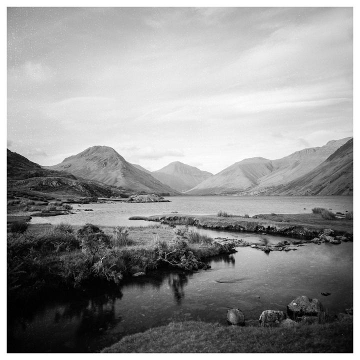 A black and white landscape shows a lake surrounded by mountains under a cloudy sky.