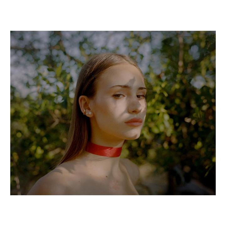 A woman with a red ribbon around her neck stands in front of green foliage, with shadows cast across her face.