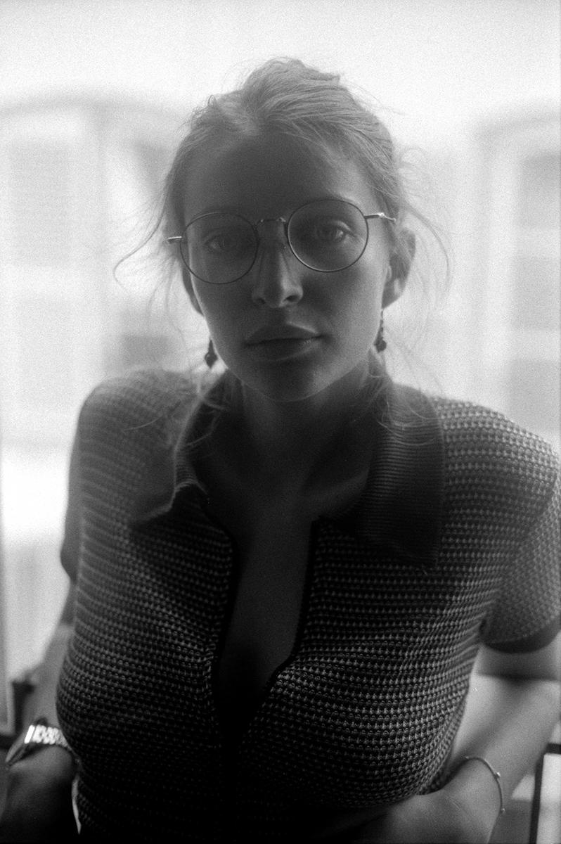 A woman wearing glasses and a patterned shirt looks directly at the viewer.