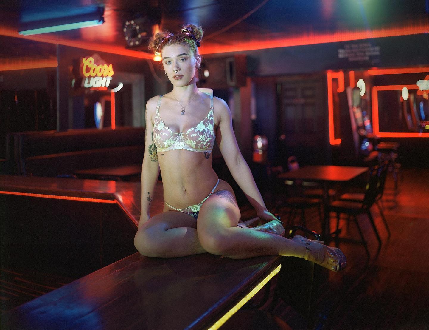 A woman in lingerie sits on a bar in a dimly lit strip club with neon signs.