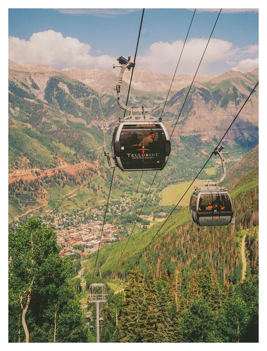 Two gondola cars are suspended by cables over a mountain valley with a town nestled among trees.
