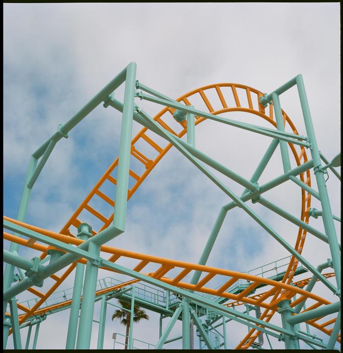 A roller coaster with teal supports and orange tracks curves against a cloudy sky, with a palm tree visible in the background.
