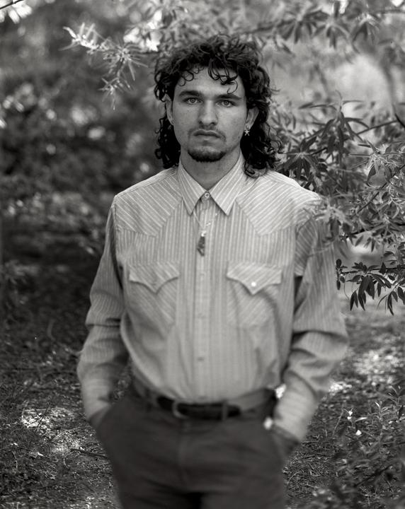 A man with curly hair and a beard stands outdoors wearing a striped shirt and a necklace.