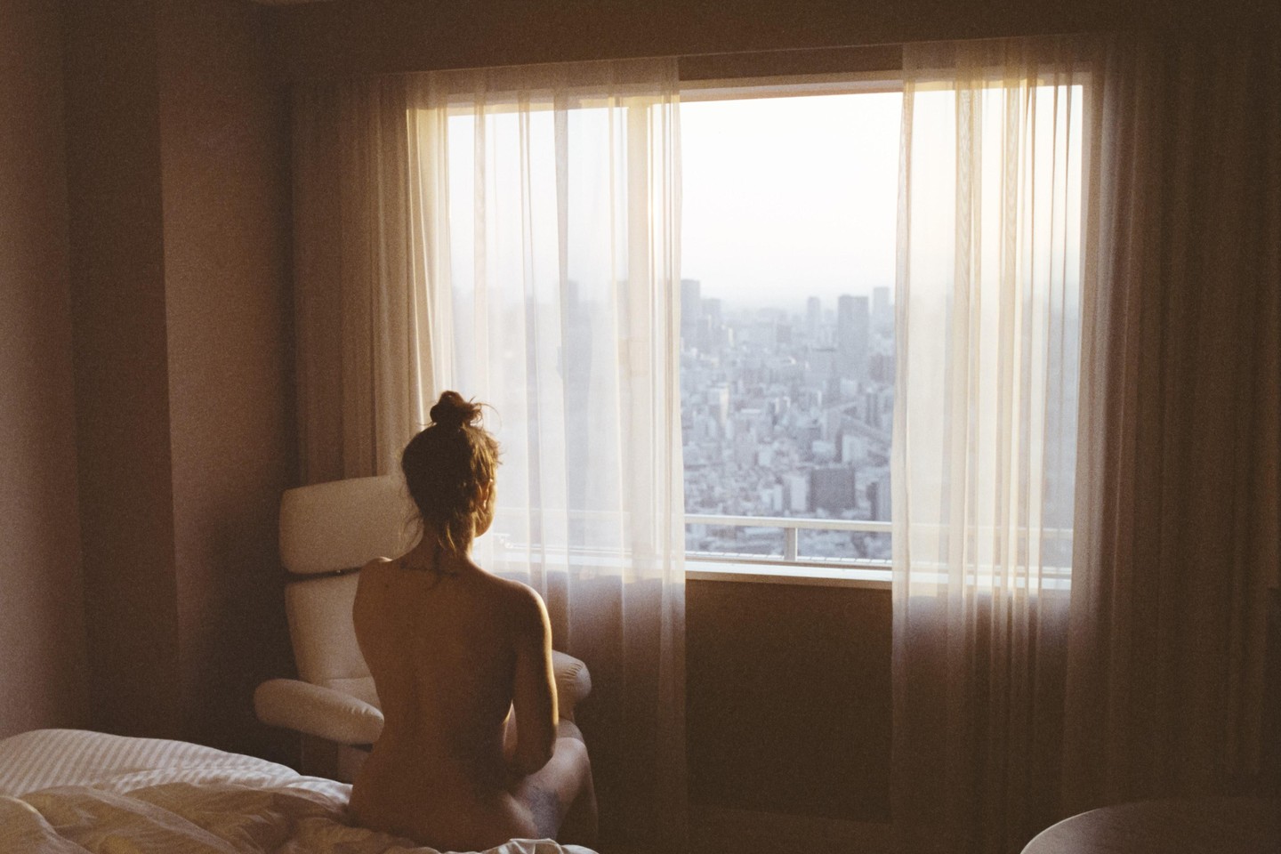 A woman sits on a bed looking out a window at a city skyline.
