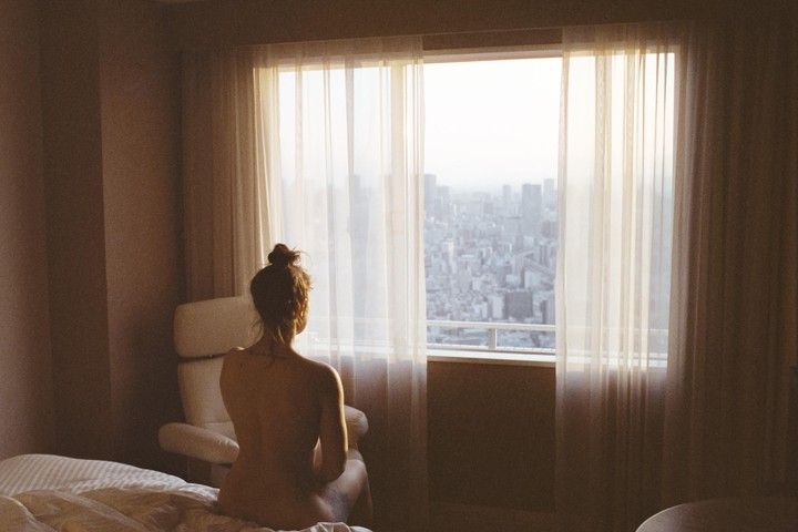 A woman sits on a bed looking out a window at a city skyline.
