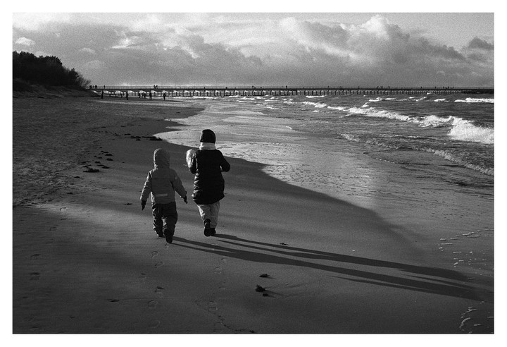Two children walk along a sandy beach with a pier in the distance and waves crashing on the shore.