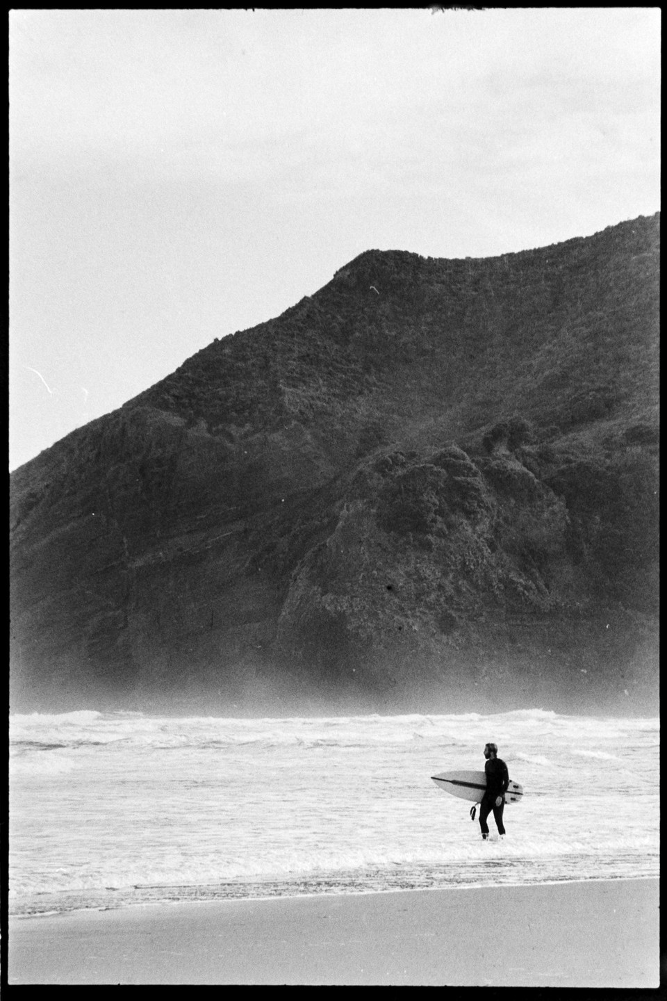 A surfer in a wetsuit walks into the ocean with a surfboard on a beach with a large mountain in the background.