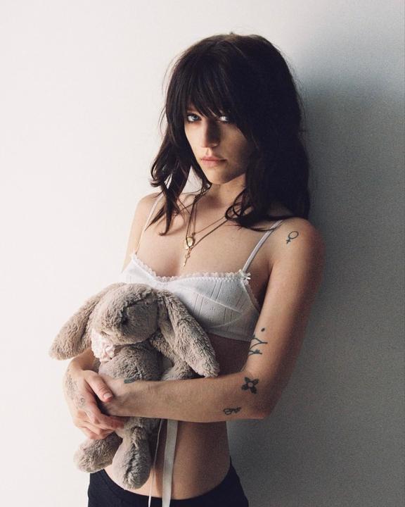 A woman with dark hair and bangs wears a white bra and holds a stuffed bunny against a white wall.