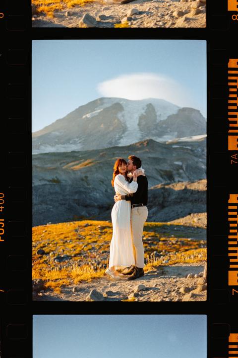 A couple embraces on a rocky, grassy hill with a large, snow-capped mountain in the background.