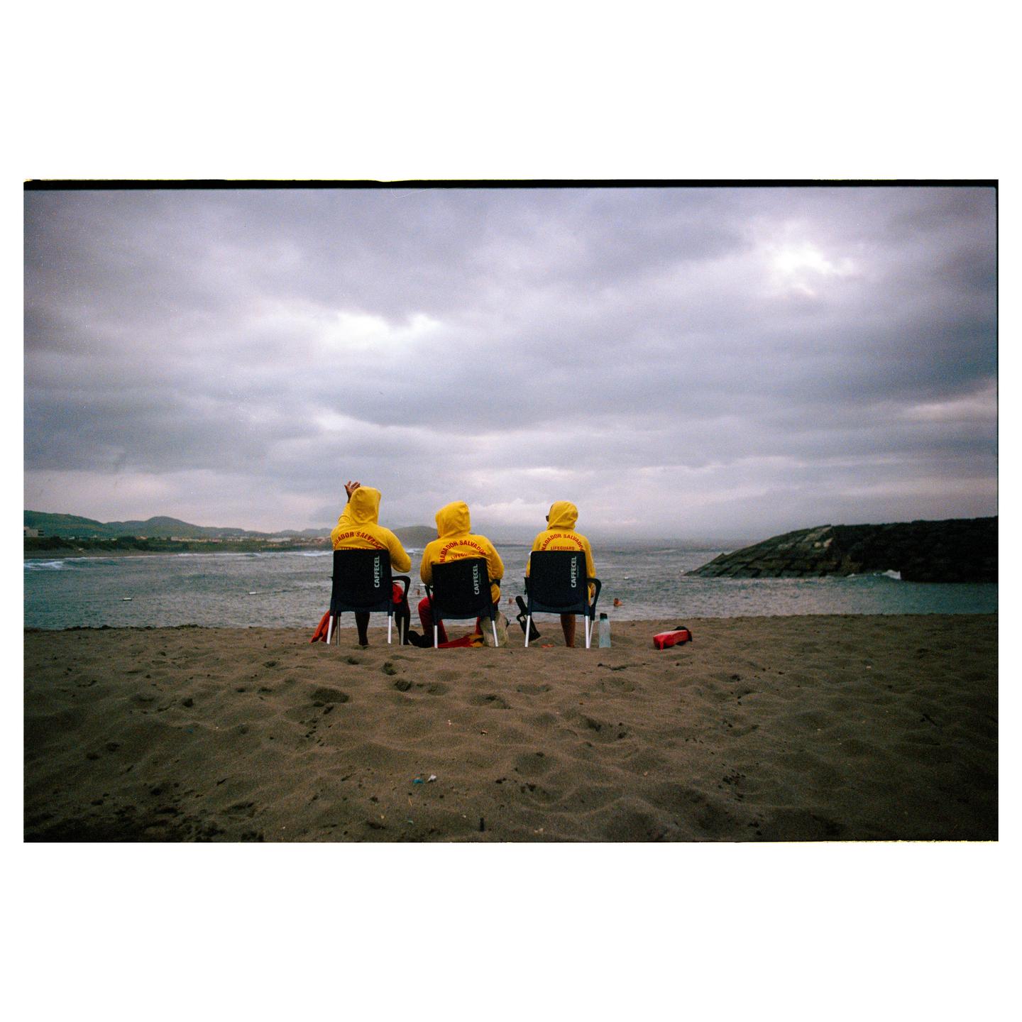 Three people in yellow hooded jackets sit in chairs on a sandy beach, looking out at the ocean under a cloudy sky.