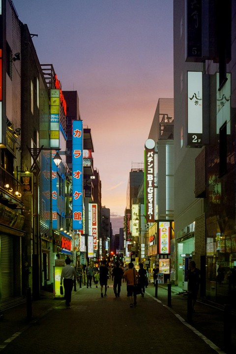 People walk along a narrow street lined with tall buildings and neon signs at dusk.