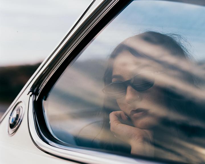 A woman wearing sunglasses is visible through the window of a car, resting her chin on her hand.