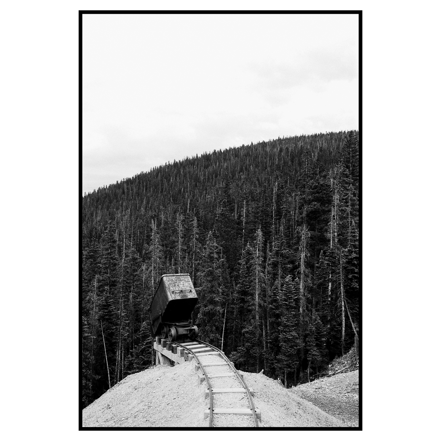 A mining cart sits on a track on a hill of dirt with a dense forest in the background.