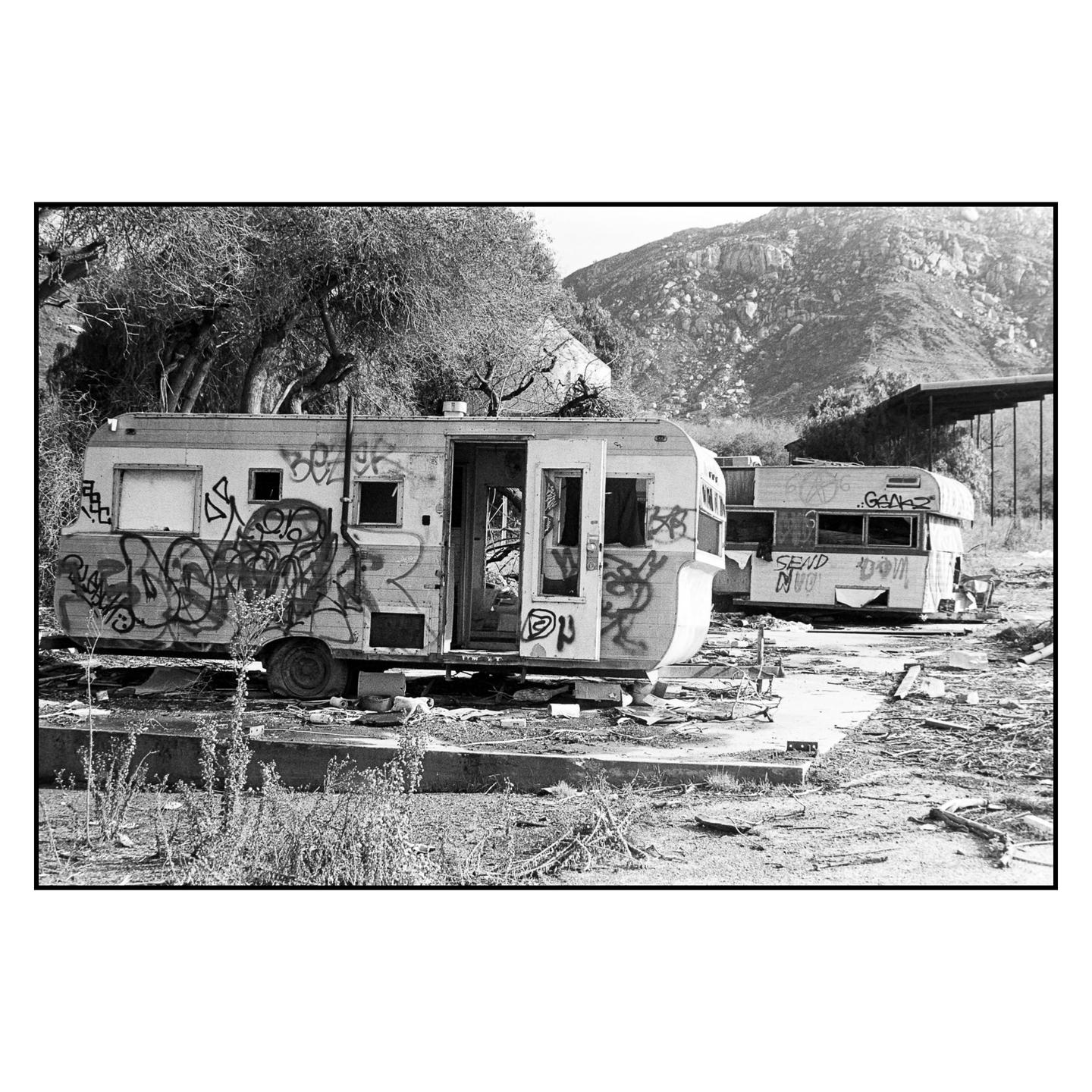 Two abandoned and vandalized recreational vehicles sit in a desolate, overgrown area with a mountain in the background.