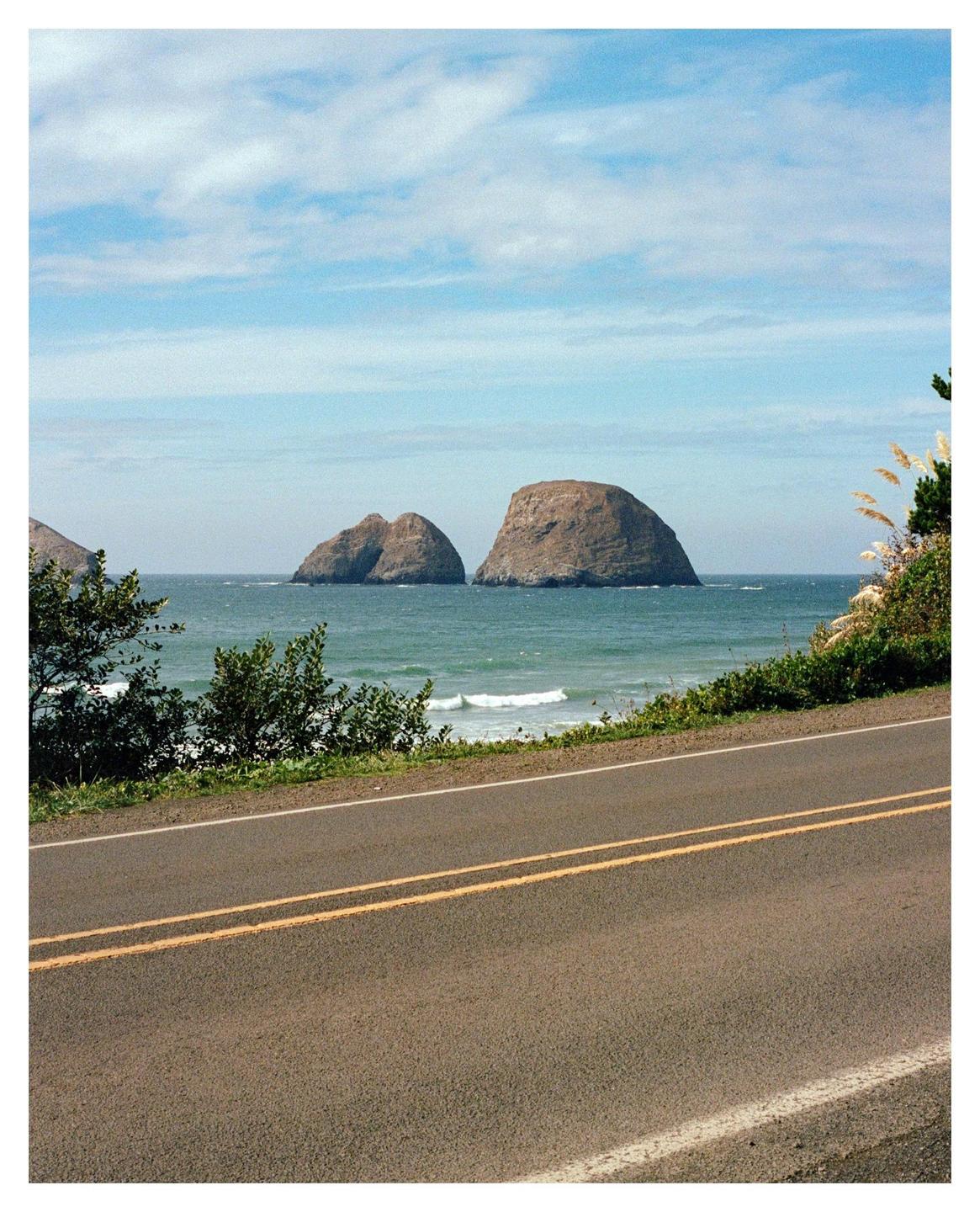 A road runs along the coast with large rock formations in the ocean under a blue sky.