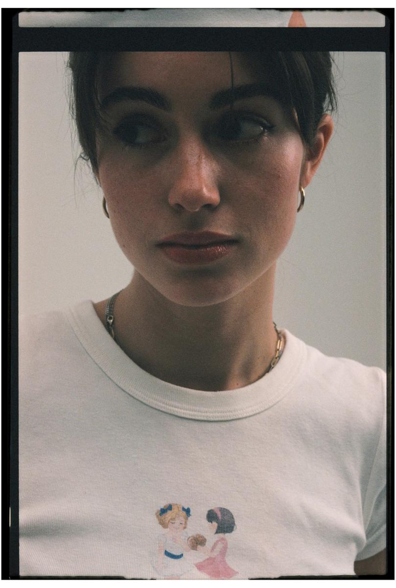 A woman with dark hair and hoop earrings looks to the side, wearing a white t-shirt with a graphic of two girls.