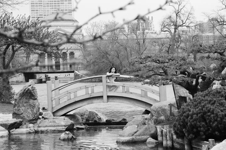 A woman leans on a wooden bridge over a pond in a park with buildings in the background.