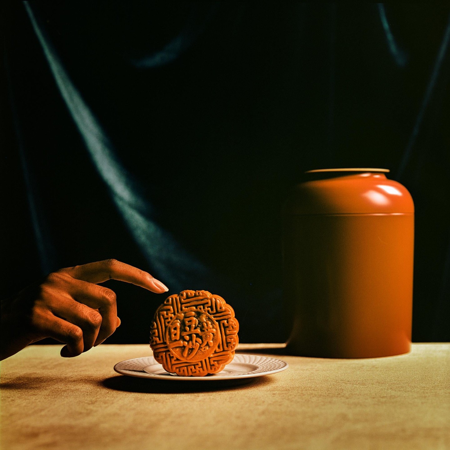 A hand points to a mooncake on a small plate next to a terracotta jar.