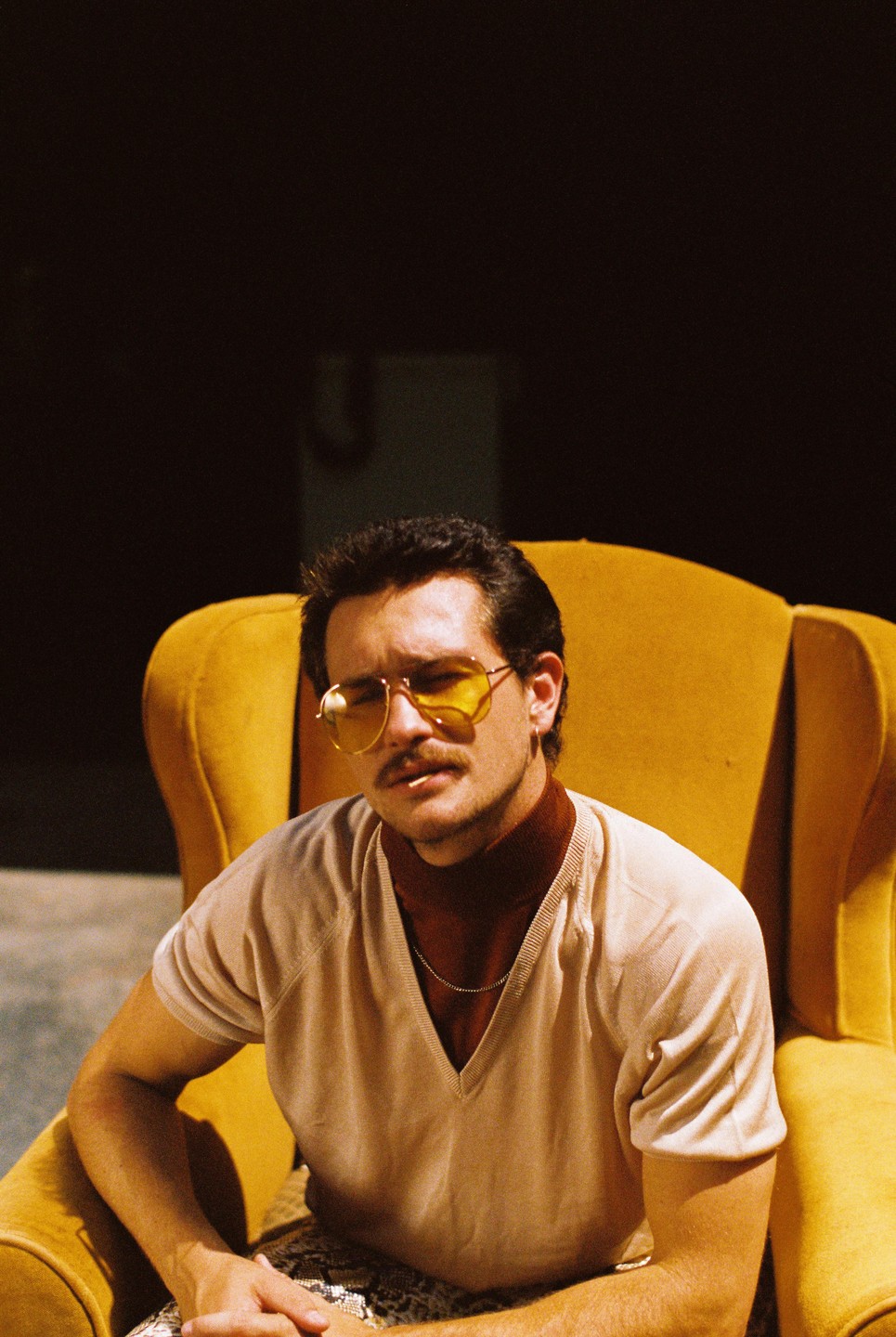 A man with a mustache and yellow-tinted sunglasses sits in a yellow armchair.