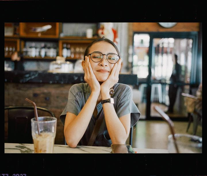 A woman with glasses rests her chin in her hands at a table with a drink.