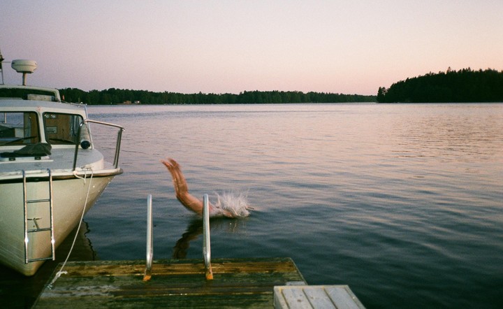 A person dives into a lake from a wooden dock next to a boat, with a forest on the far bank under a pale sky.