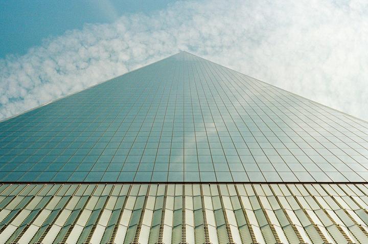 The glass facade of a skyscraper reflects the sky and clouds, with the building tapering towards a point.