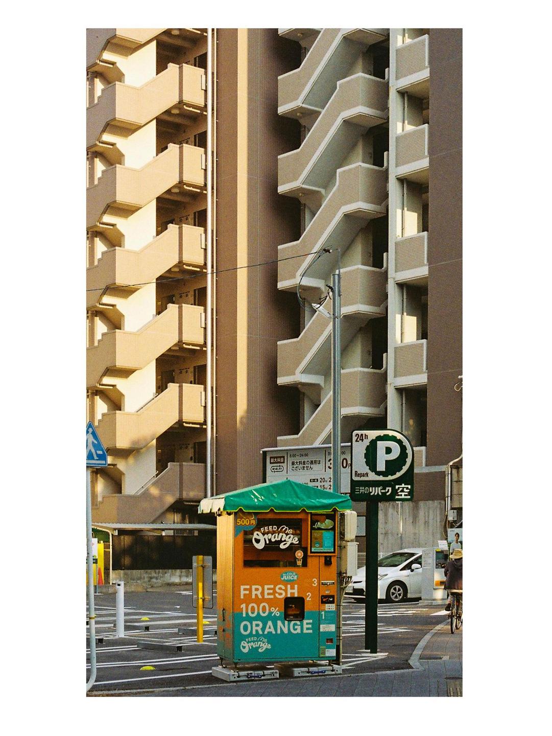 An orange juice vending machine stands on a city street with apartment buildings in the background.